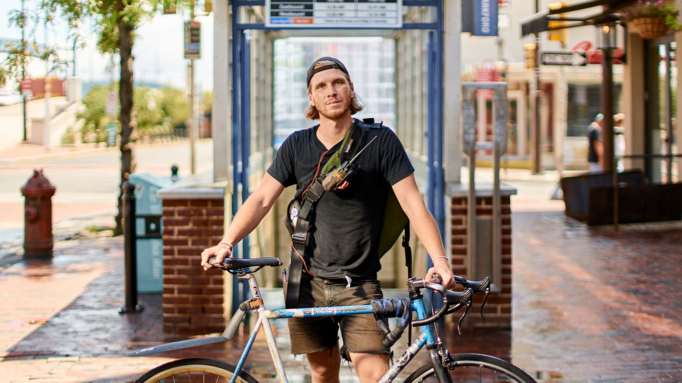 A young man with blonde hair and a hat straddles his bike and looks into the camera
